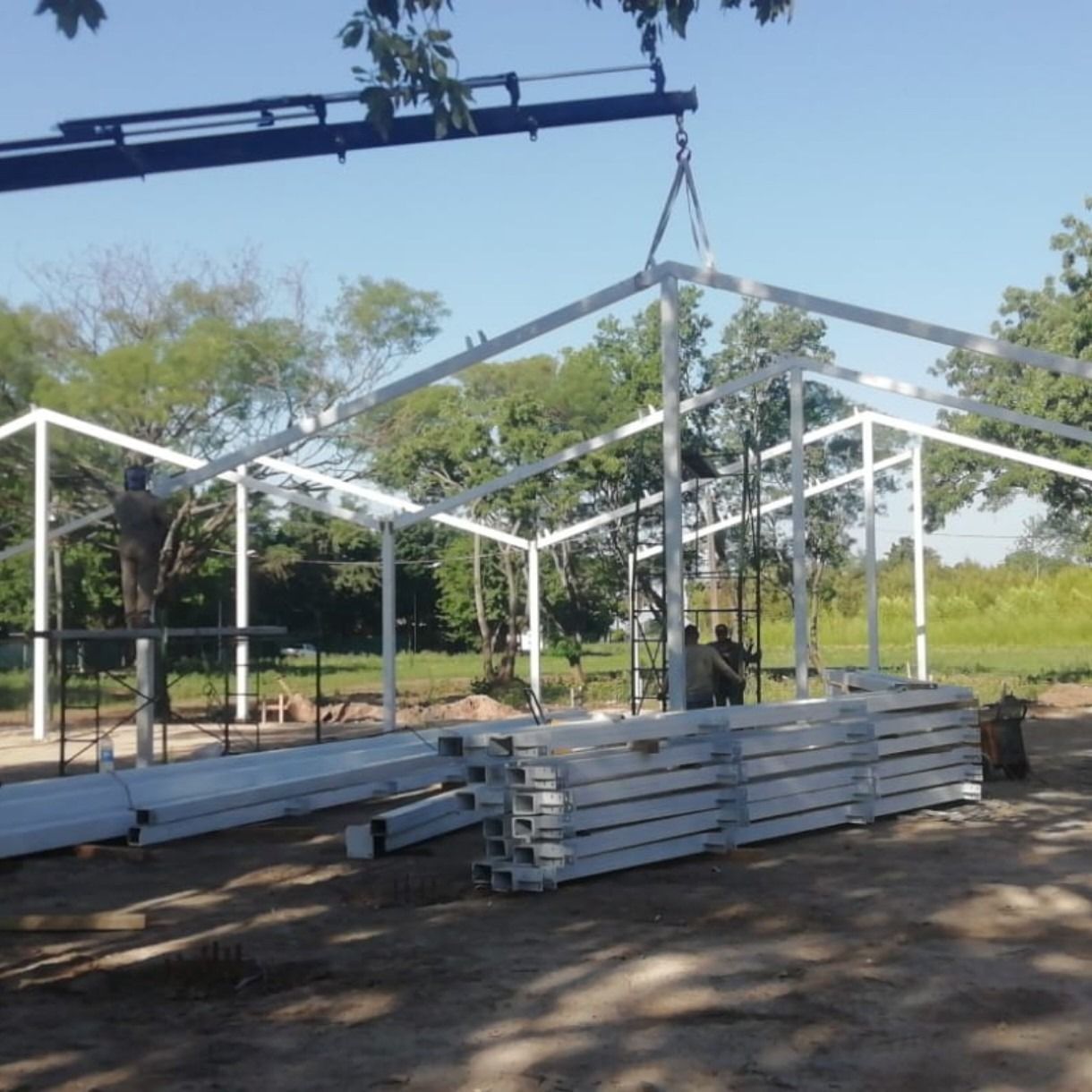 Una grúa eleva secciones de estructura metálica blanca en una obra de construcción al aire libre bajo un cielo azul.