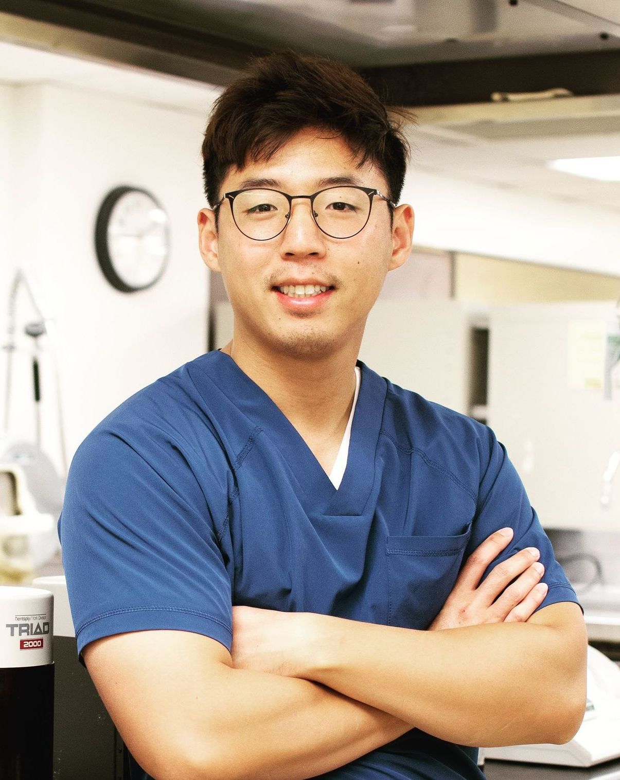 Person in blue scrubs with glasses stands arms crossed in a bright clinical setting.
