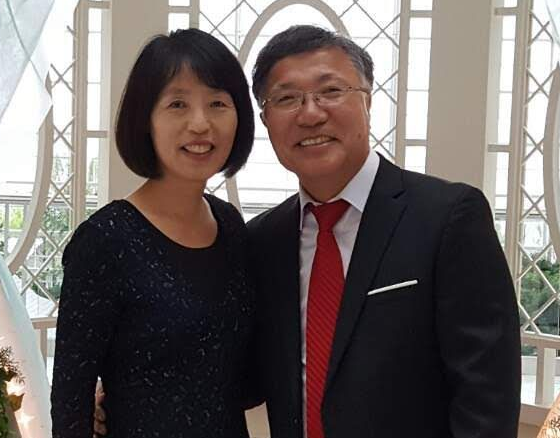 Two smiling people in formal attire standing together indoors, with a bright windowed background.