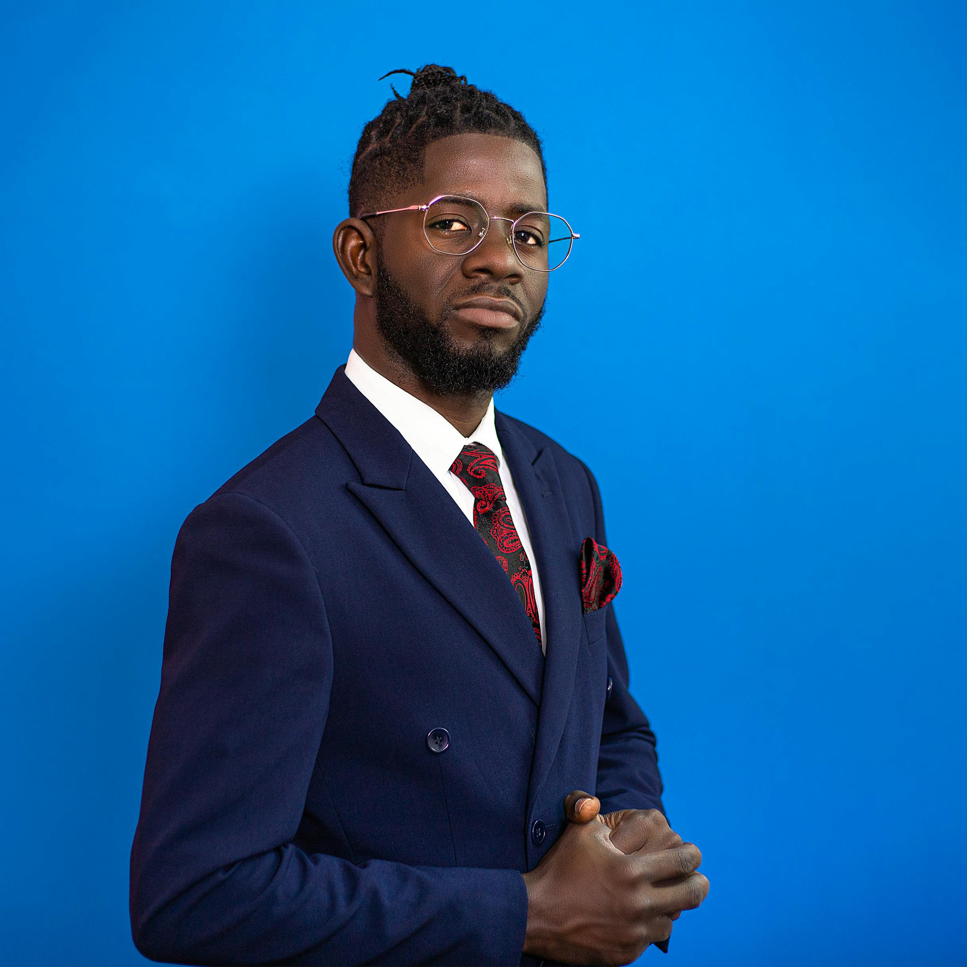 Man in a suit, eyeglasses, and red tie, arms crossed, poses against a blue background.