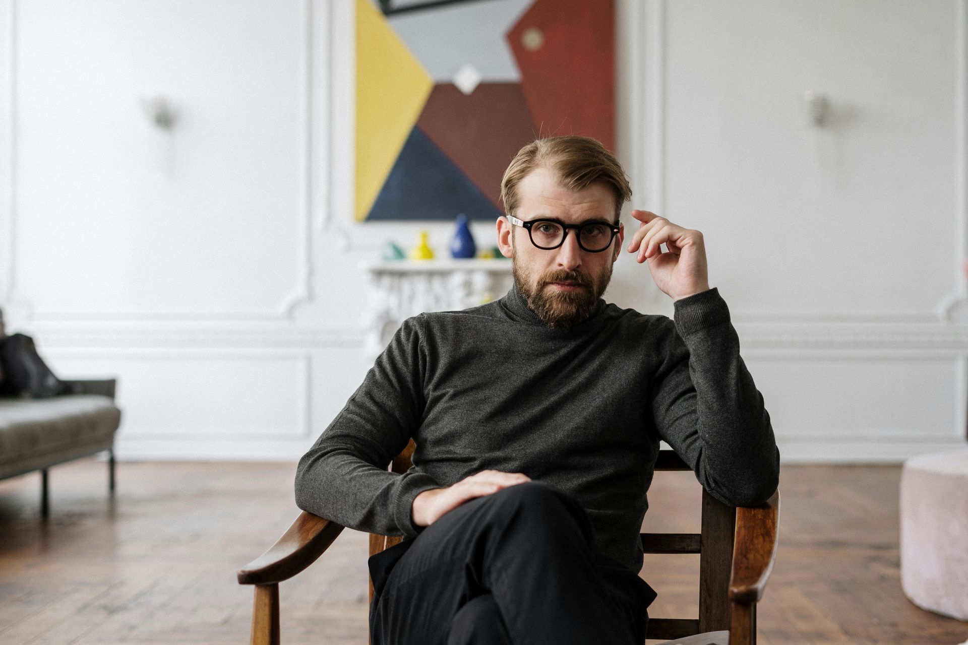 Man in glasses sits, hand to face, in chair; abstract art and decor in background.