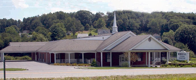 Low, brick church building with a white steeple, surrounded by trees and a gravel parking lot