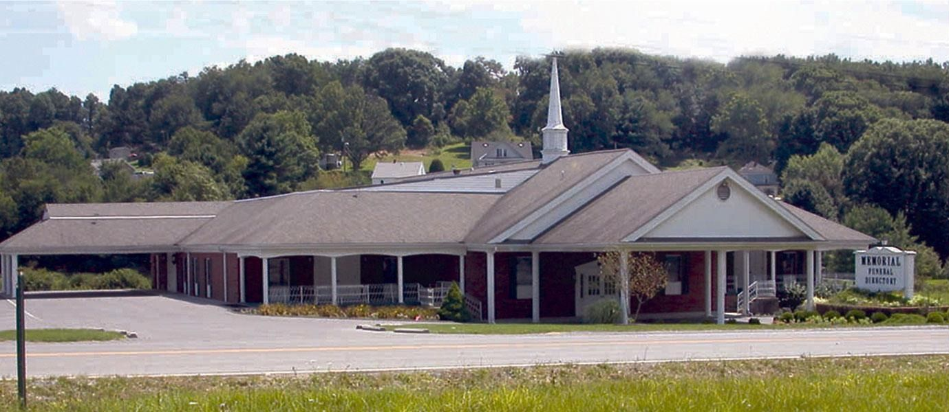 Single-story church building with a white steeple and parking lot, set against a wooded hillside.