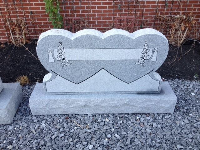 Gray heart-shaped memorial stone on a concrete base against a brick wall, with small angel figures