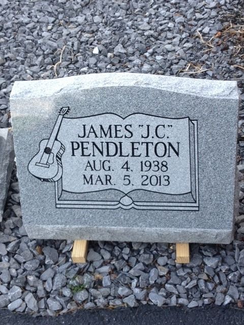 Gray gravestone for James “J.C.” Pendleton with guitar engraving, resting on wooden supports over gravel.