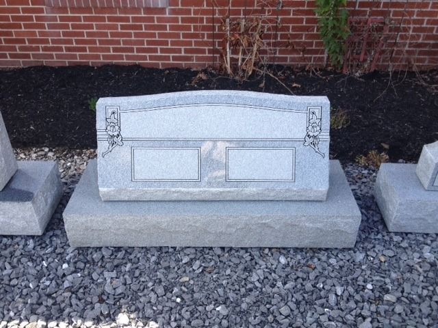 Gray engraved granite tombstone in a cemetery, set on gravel with a brick wall behind it