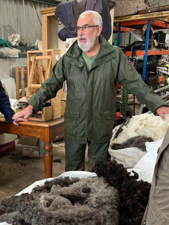 Man in green jacket speaking in a workshop, standing beside tables with piles of wool and tools