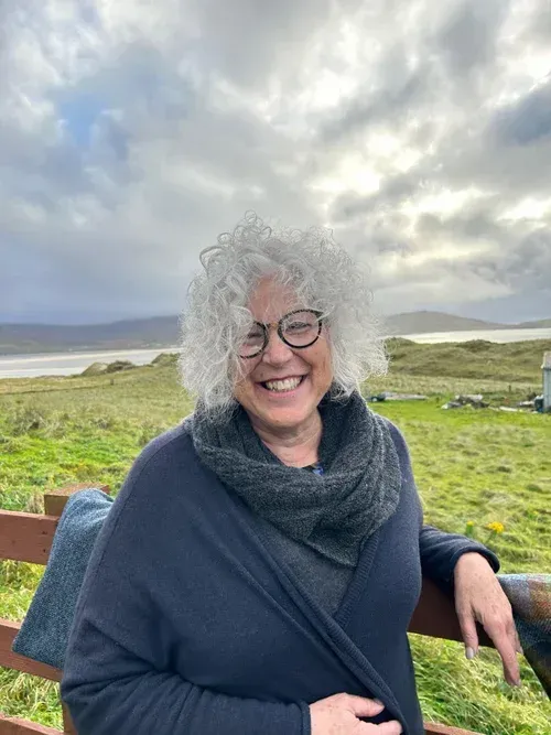 Smiling person with curly gray hair and glasses in a navy wrap, standing by a bench in a grassy coastal landscape