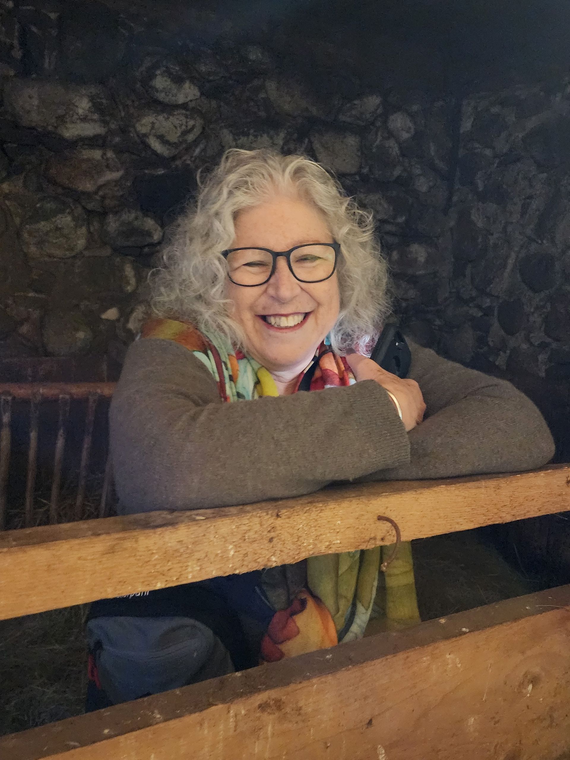 Smiling person with glasses leaning on a wooden railing in a dimly lit outdoor setting