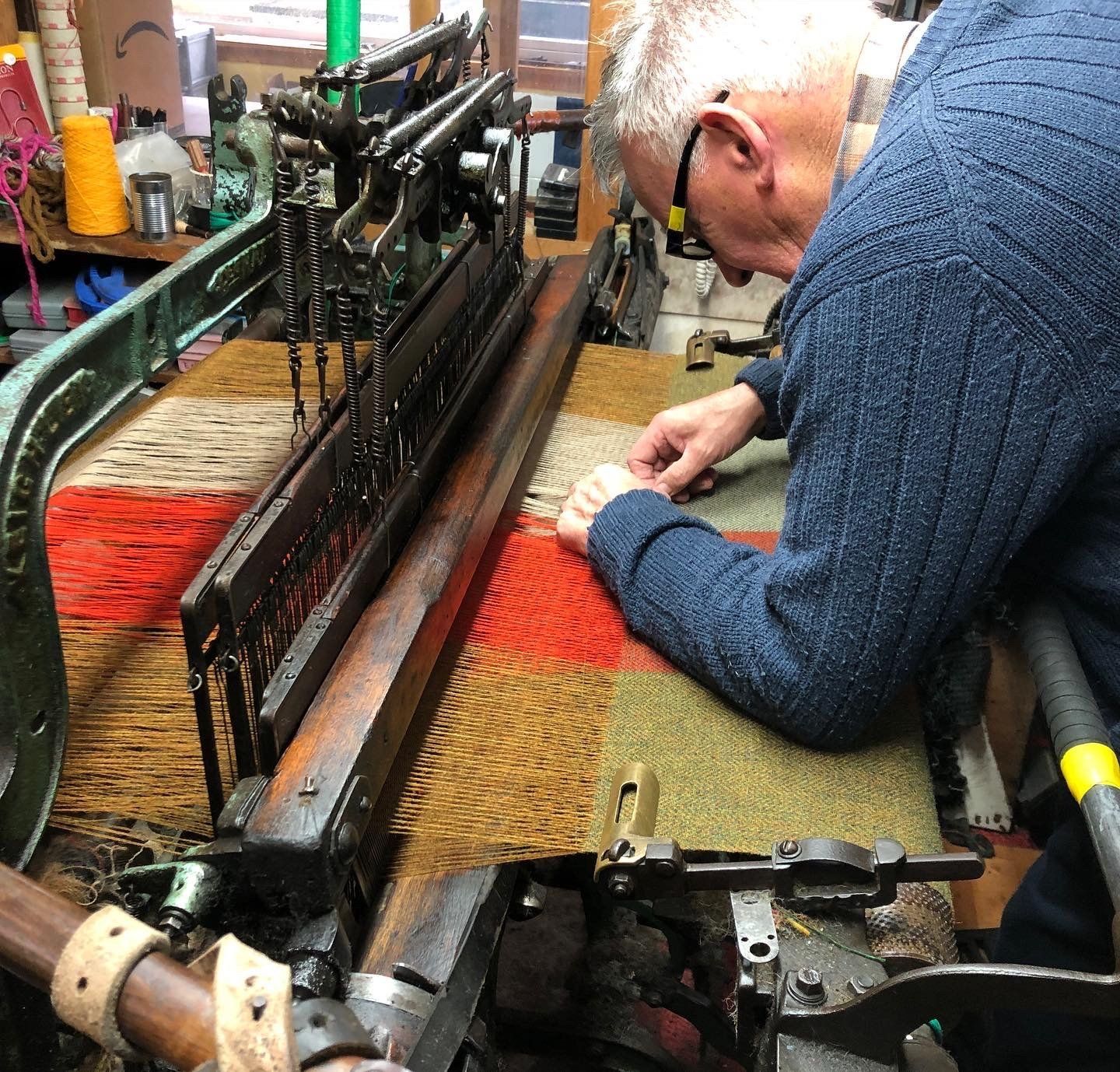 Man working on a loom, weaving red, tan, and orange fabric in a workshop.