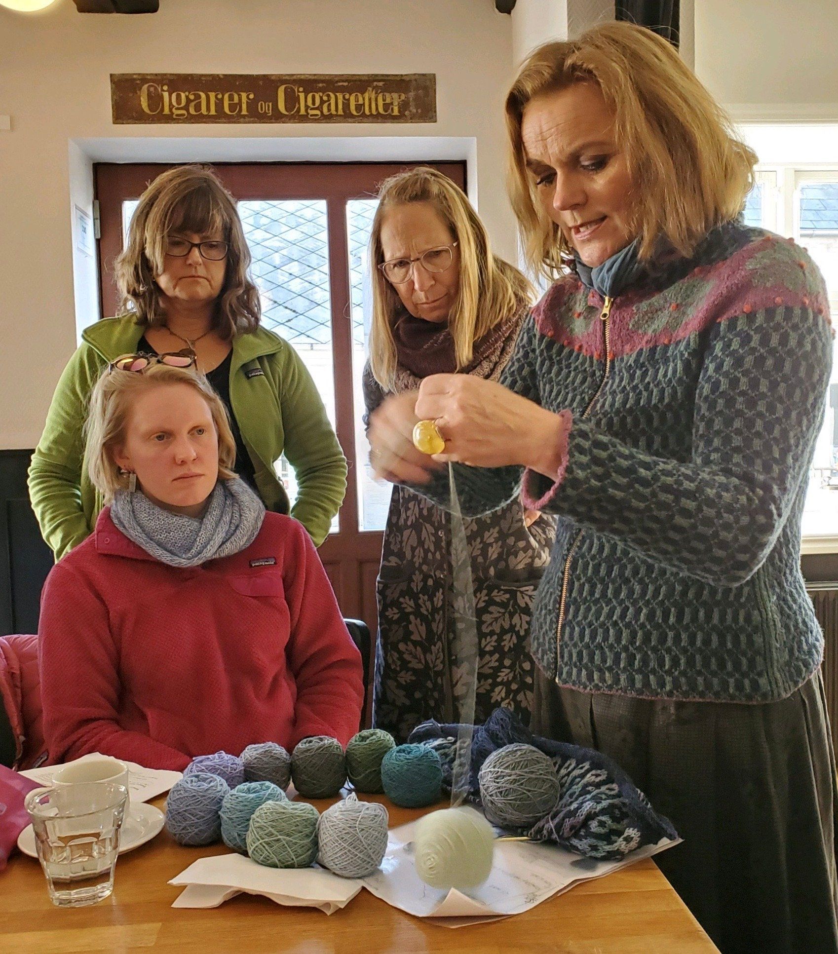 Women gather around a table, one crocheting yarn balls while others watch in a craft room.