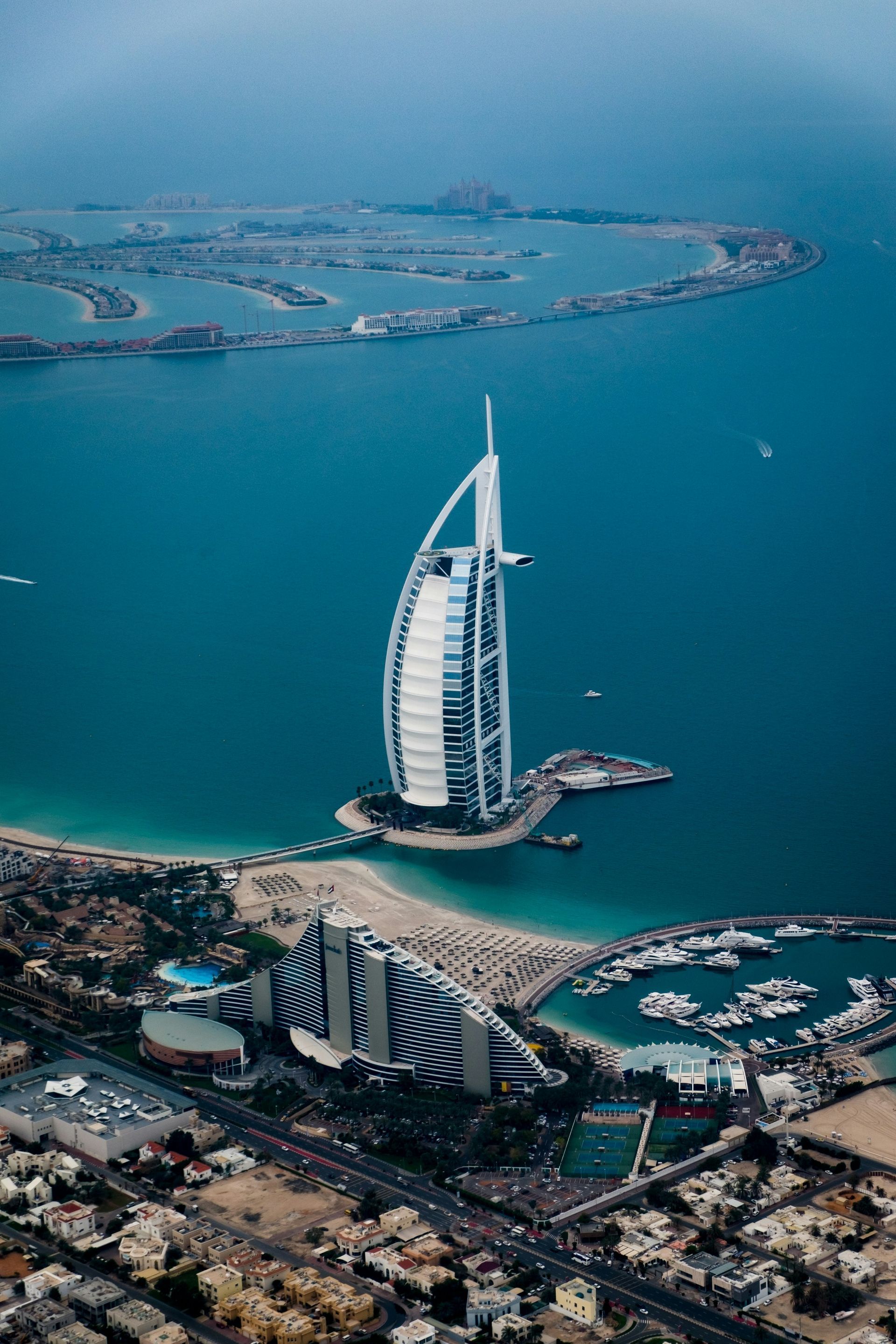 Aerial view of Dubai’s Burj Al Arab on a turquoise coastline with a marina and nearby buildings