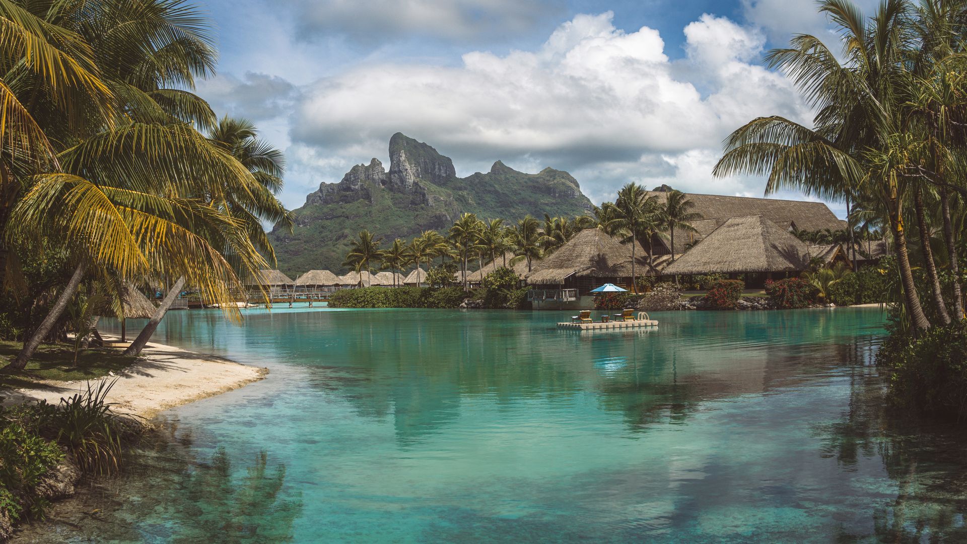 Tropical lagoon with turquoise water, palm trees, sandy beach, and huts beneath dramatic mountains and clouds