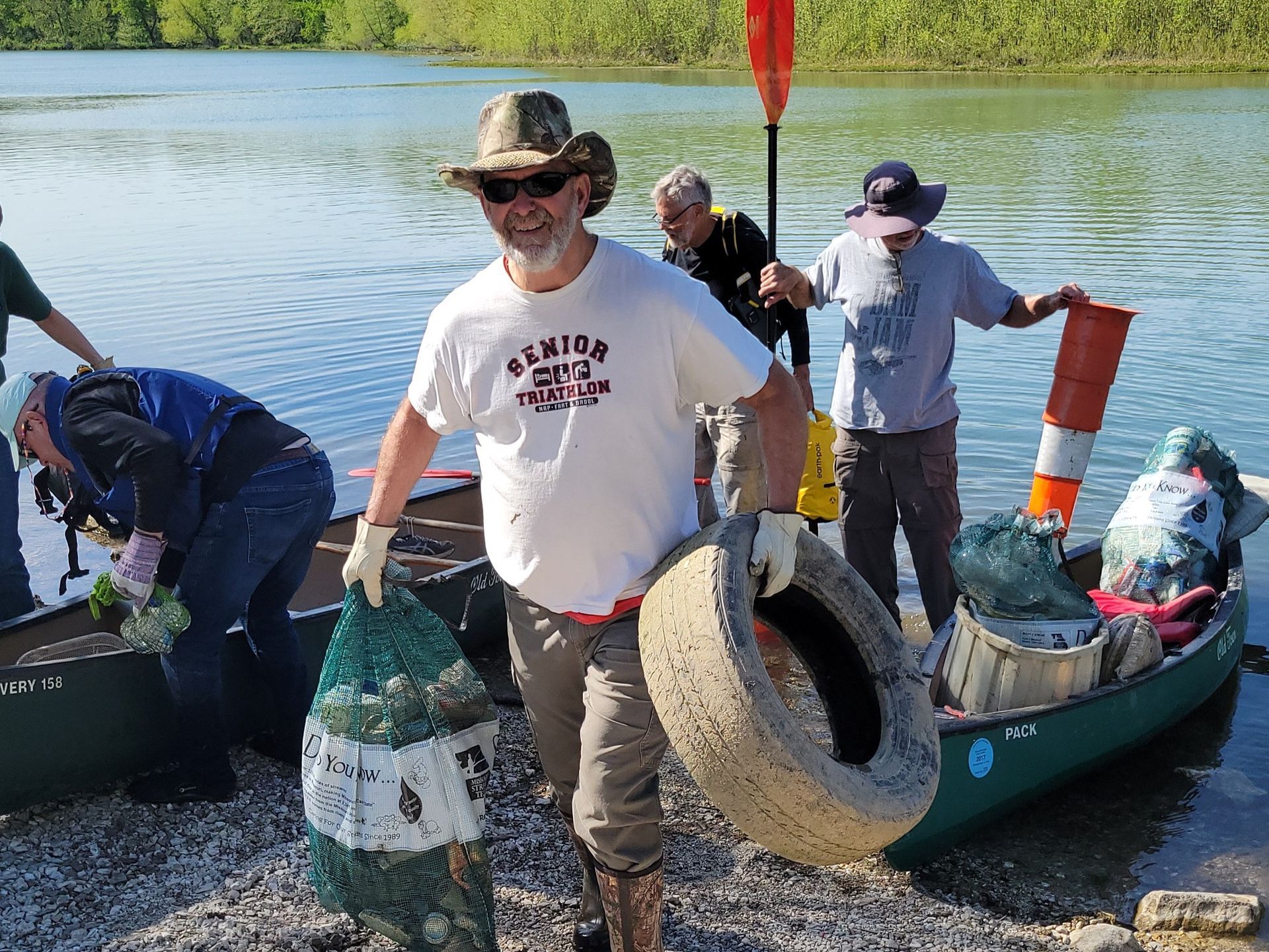 People collecting trash from a canoe beside a calm lake, with one person holding a tire.