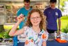 Girl holding up a small object at a science fair table, with two people and posters in the background