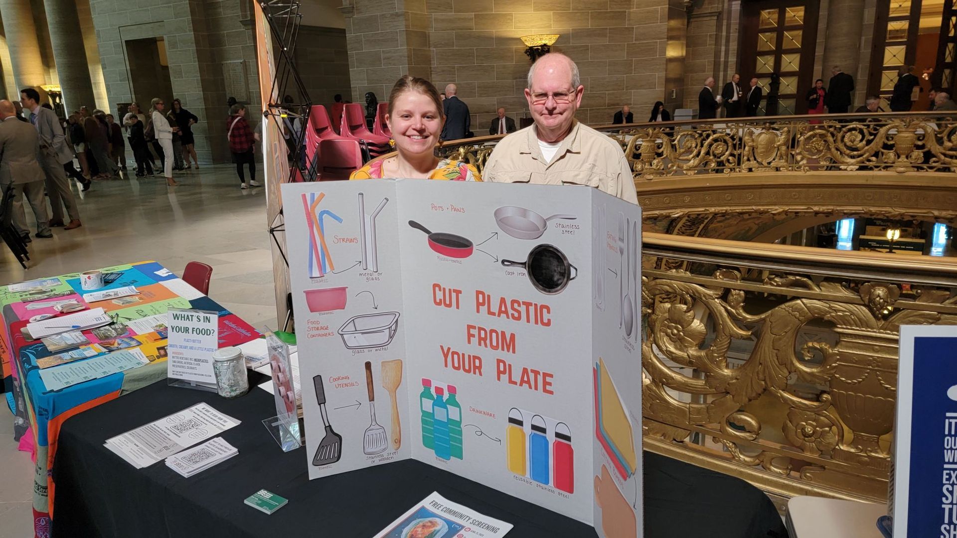 Two people hold a sign at an indoor protest table reading “Out plastic for plate” with drawings and bottles.