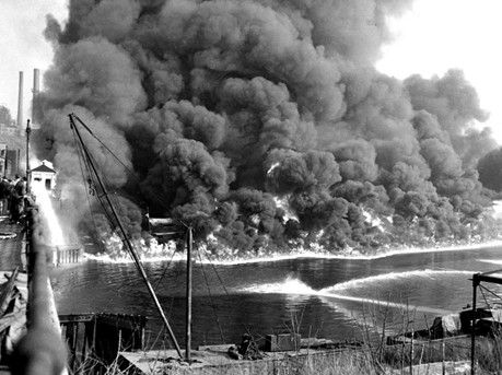 Massive fire with thick smoke billowing over water near a dock and crane