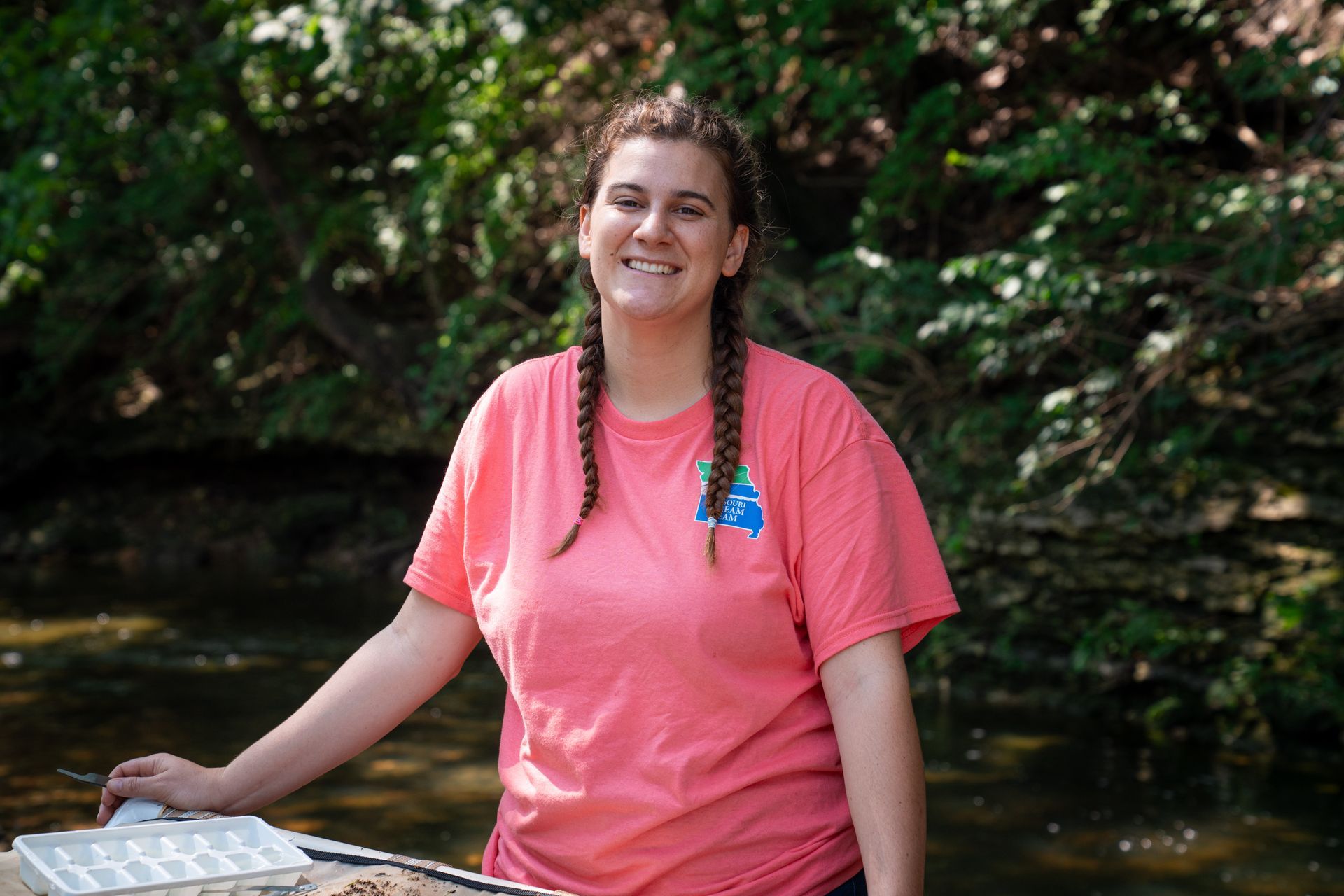 Smiling woman in a pink shirt standing by a creek, with trees in the background.