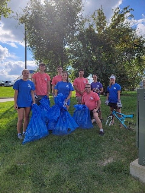 Group of volunteers in colorful shirts holding blue trash bags outdoors on a grassy lawn.