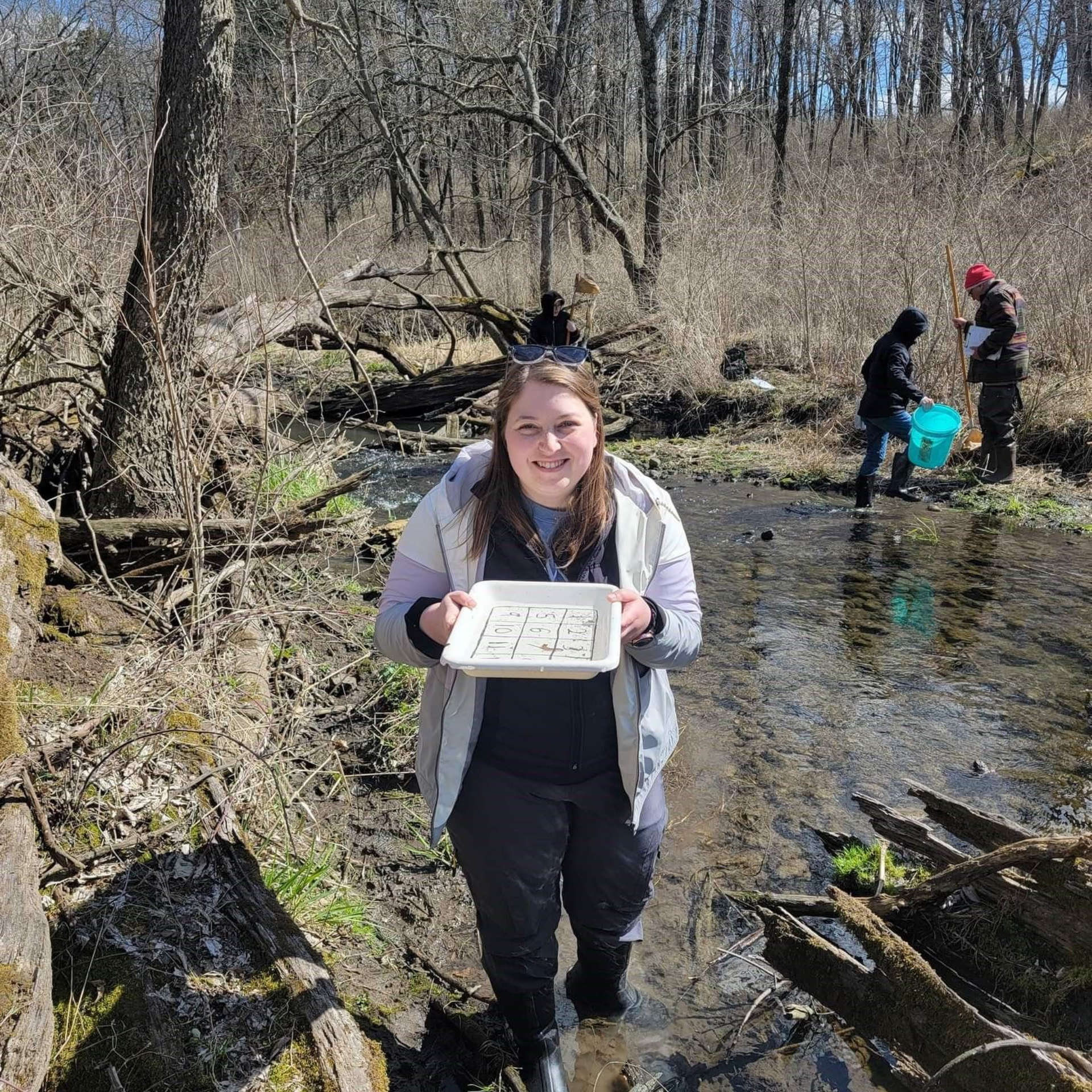Person holding a tray while standing in a shallow creek with two people in the background.