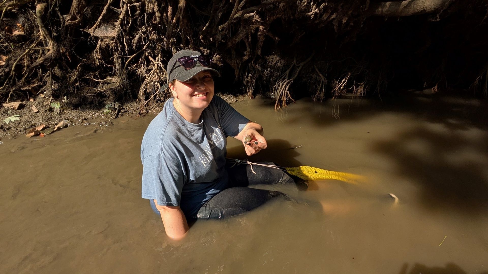 Person smiling while sitting in muddy water at a rocky stream, holding a yellow object