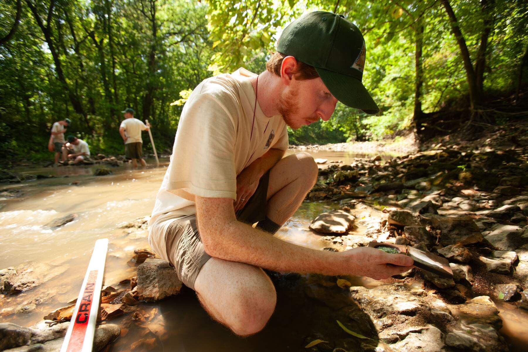 Person crouching by a muddy creek in a wooded area, examining rocks near the water