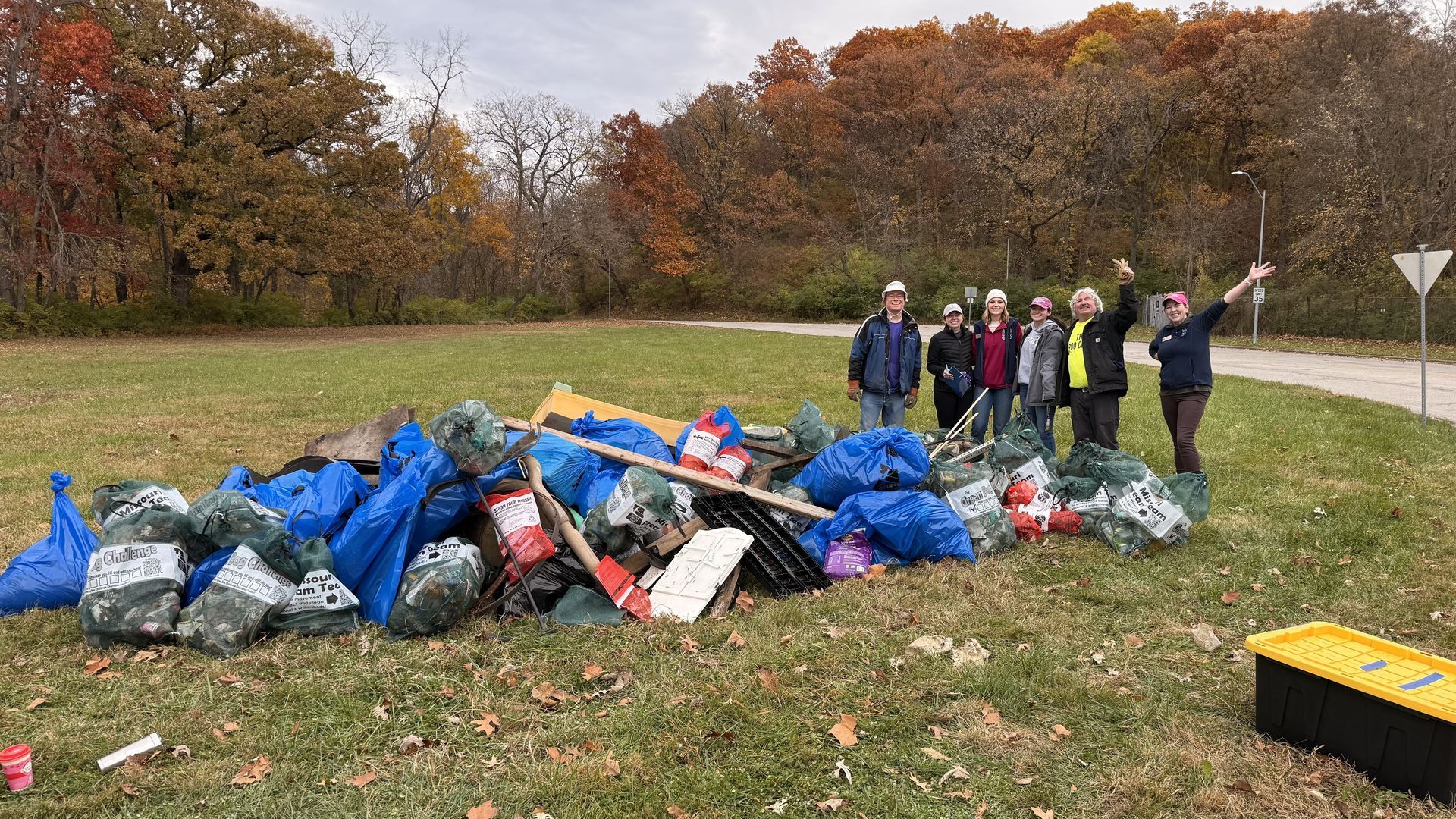 People standing by a large pile of blue trash bags in a grassy park with autumn trees.