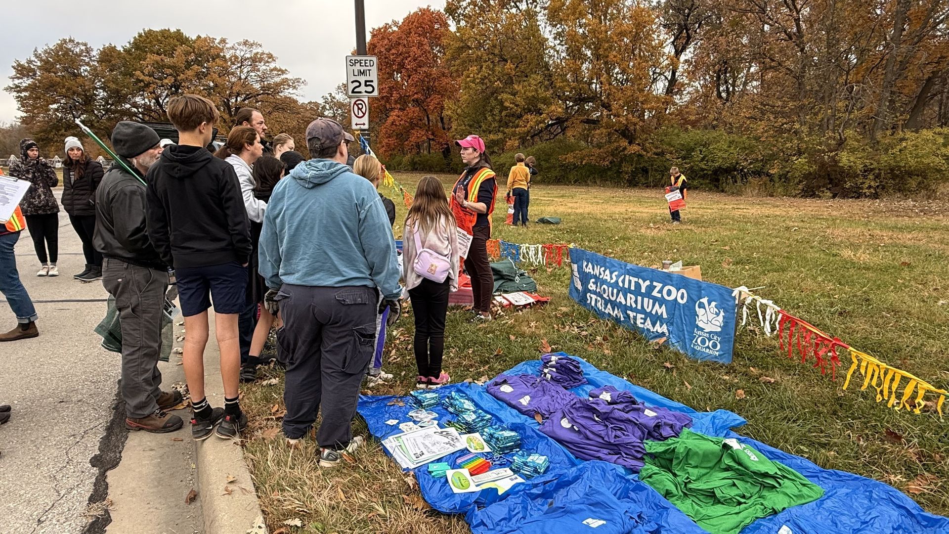People gathered beside a roadside memorial with blue signs, flowers, and colorful blankets in a grassy park area.