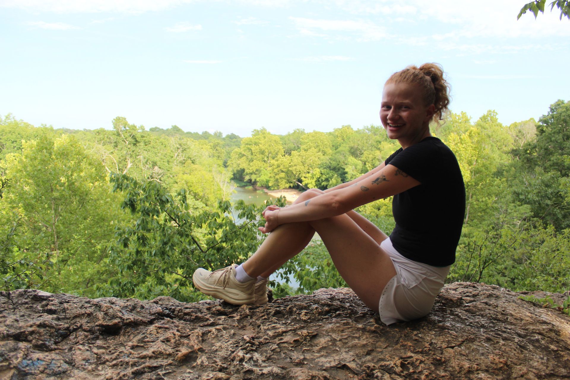 Person sitting on a rocky ledge with a forested landscape in the background, smiling in the sun.