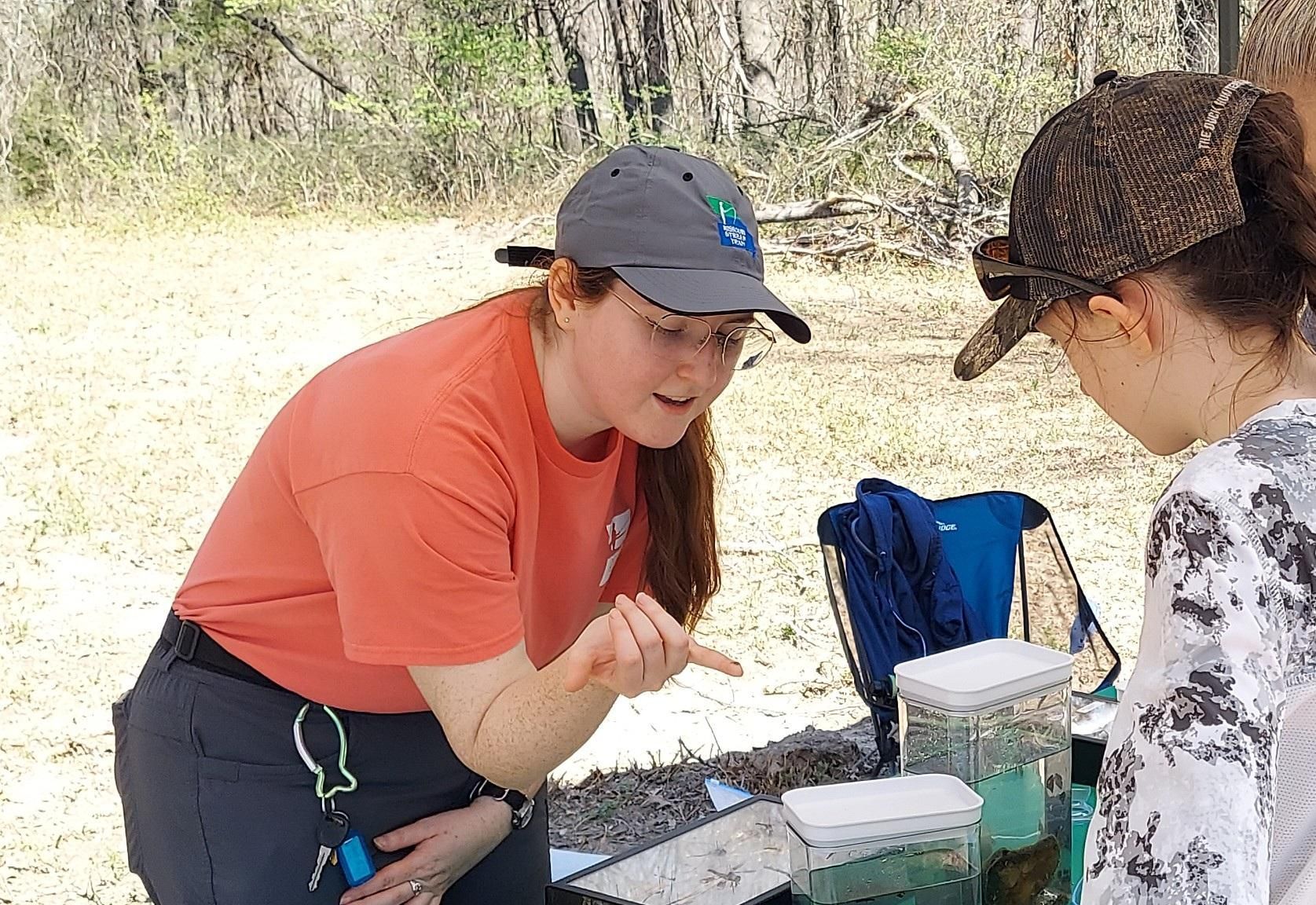 Two people examine items on a table outdoors, with a blue folding chair nearby.
