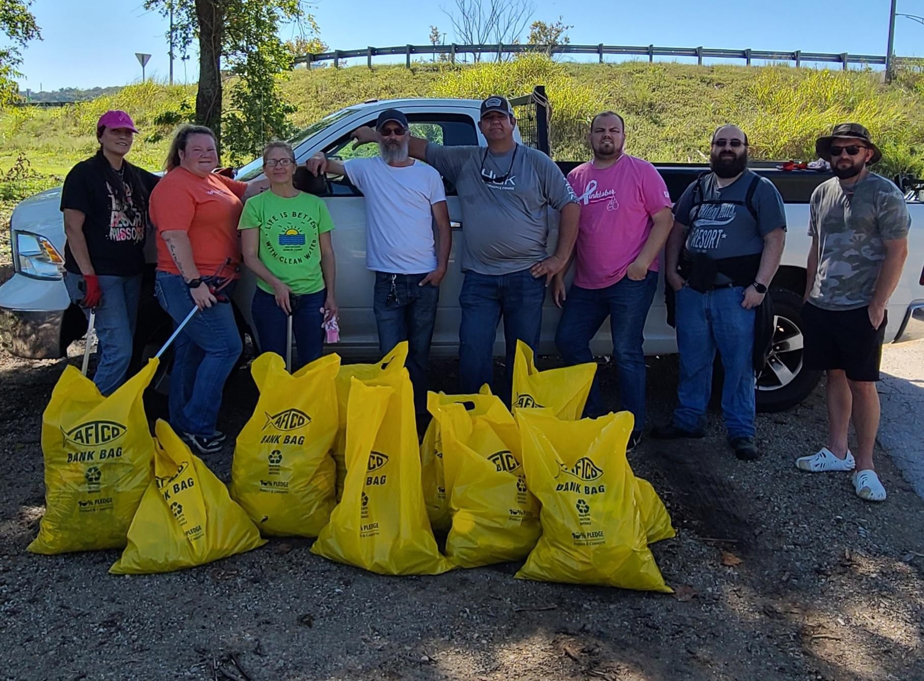 Group of volunteers posing outdoors behind yellow trash bags during a cleanup effort.