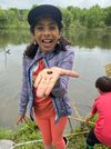 Smiling child in a cap shows a tiny frog near a pond, with another child crouching by the water.