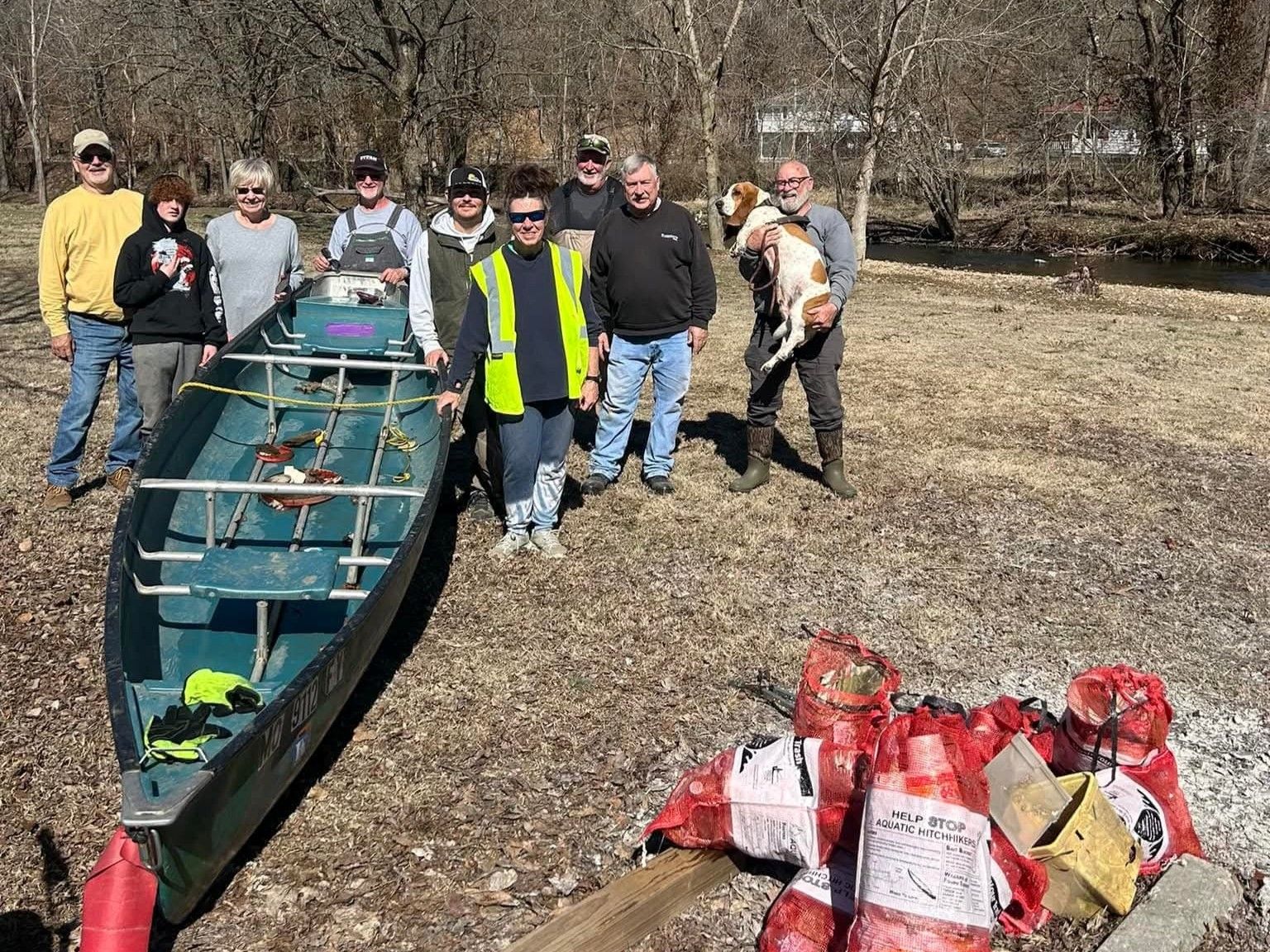 Group posing beside a canoe loaded with gear on a wooded trail, with bags and supplies in front.