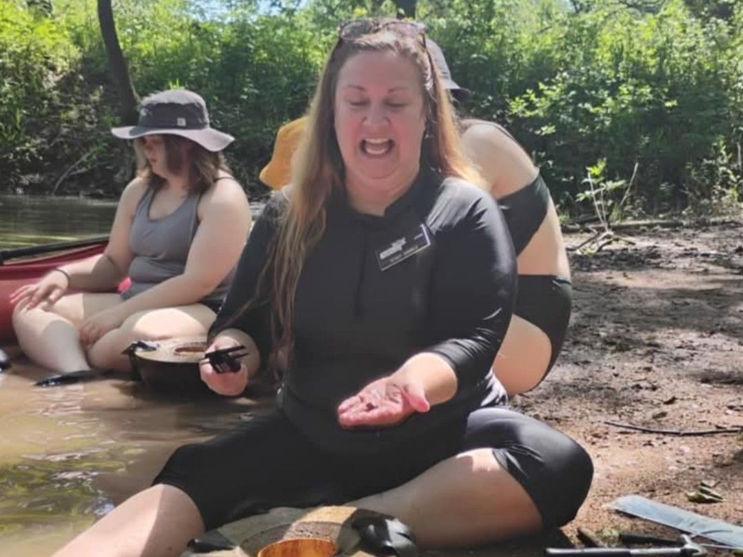 Group sitting in a muddy creek, one woman smiling and holding out her hands in a black wetsuit.