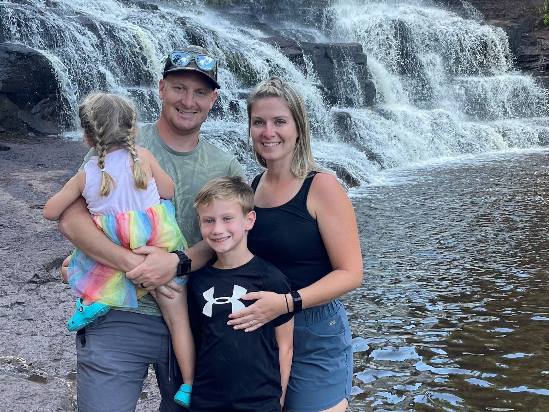 Family posing in front of a waterfall, smiling in shallow water on rocks.
