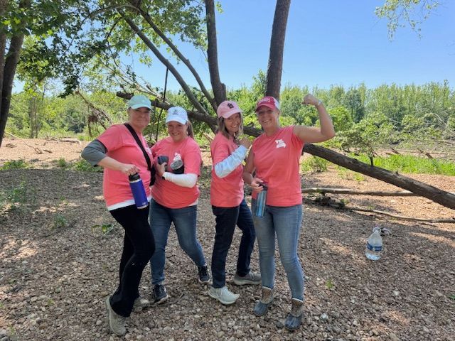 Four hikers in pink shirts and hats posing on a rocky trail under trees on a sunny day