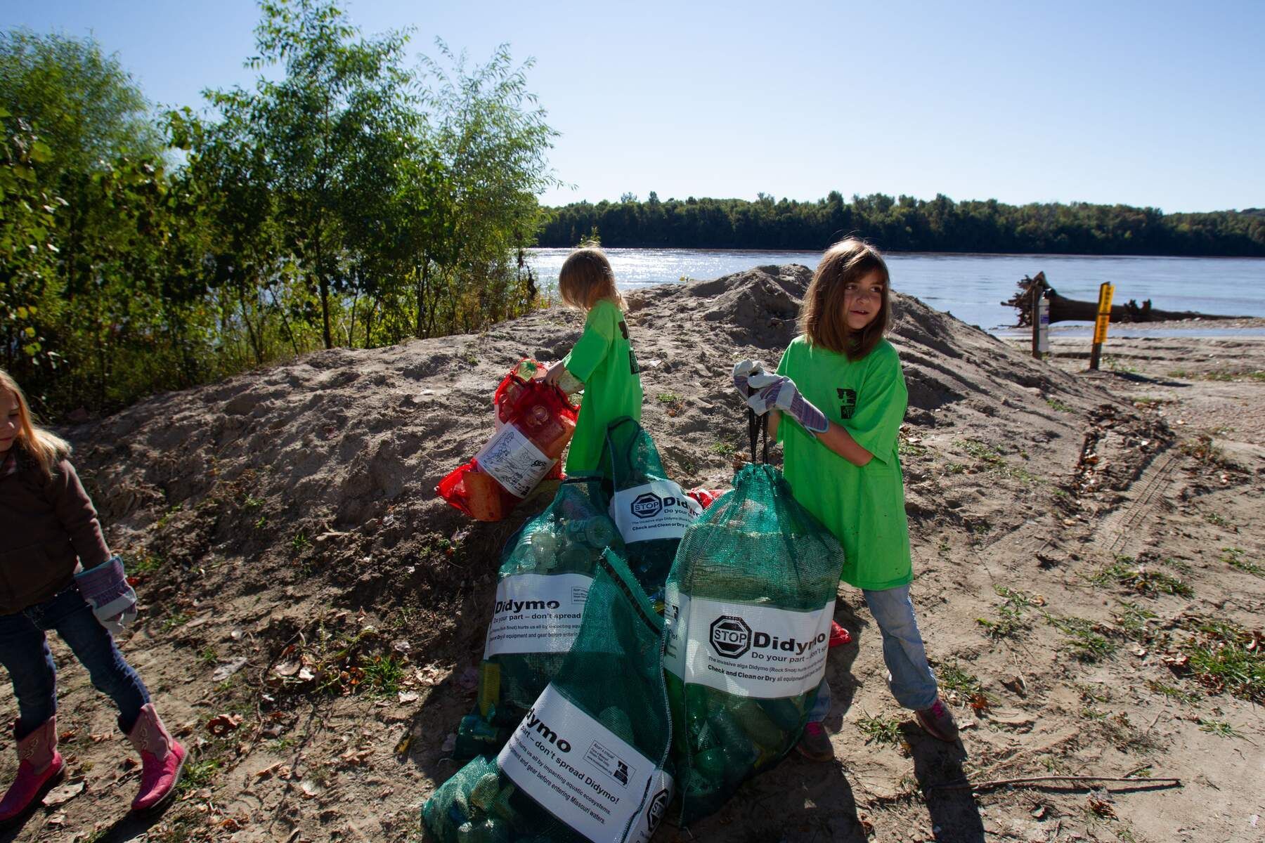 People in green shirts collecting trash on a sandy lakeshore near the water