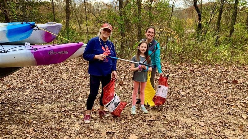 Three people stand by colorful kayaks in a wooded area, holding trash bags during a cleanup.