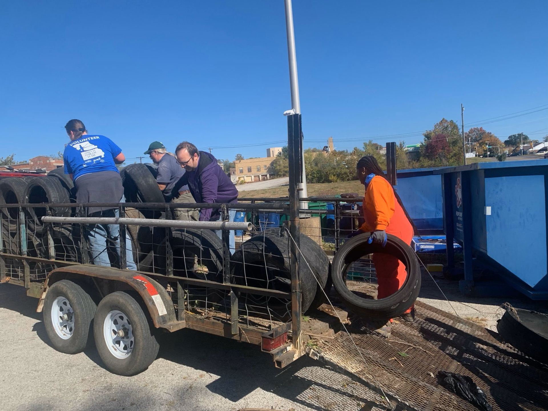 Workers loading debris onto a trailer beside a blue dumpster on a sunny day