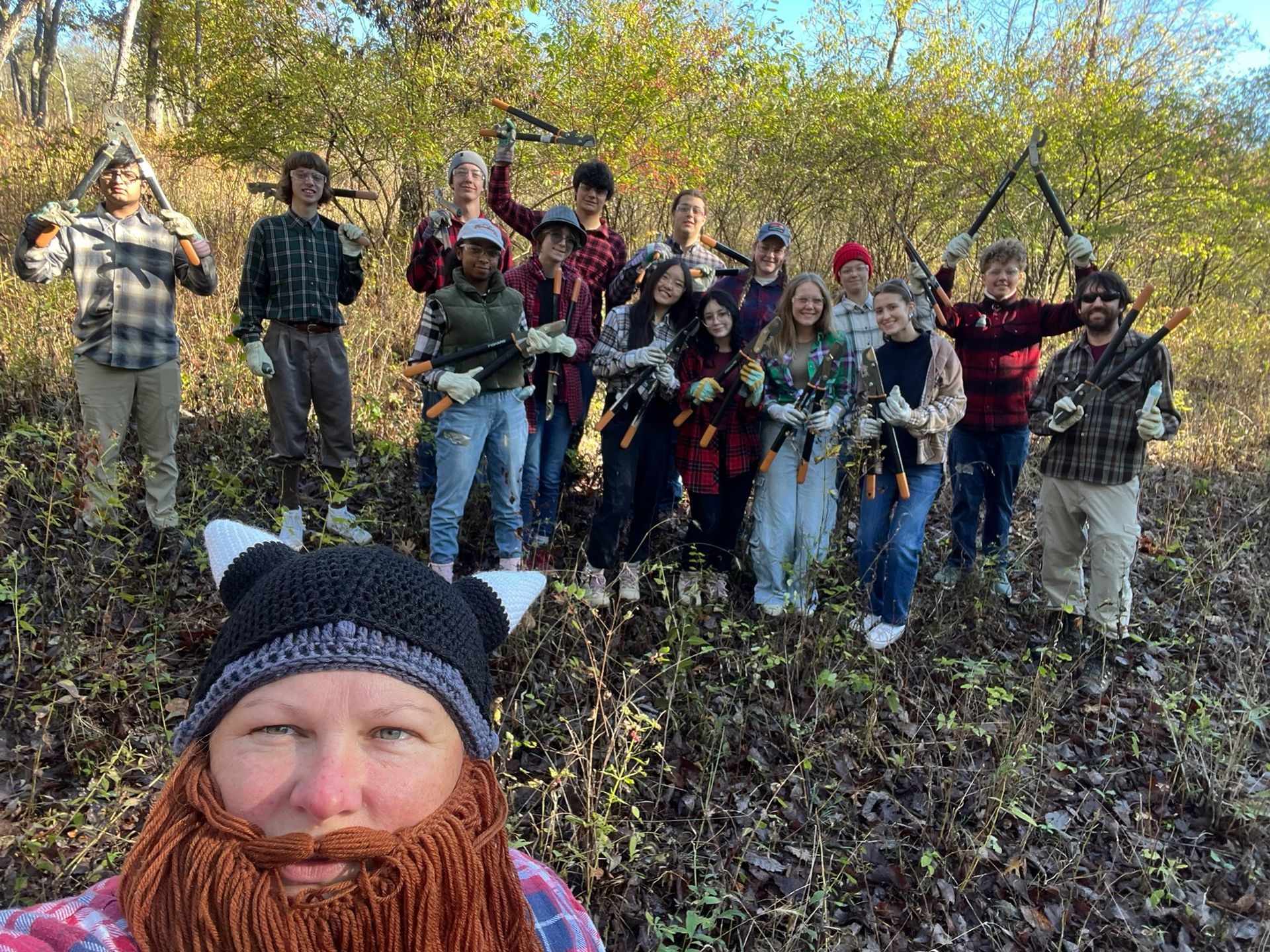 Group of people outdoors in a wooded area, smiling and raising tools or branches, with a bearded person in front.