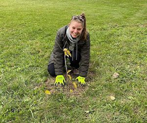 A woman is kneeling down in the grass planting a tree.