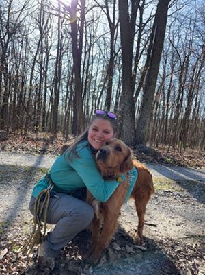 A woman is kneeling down next to a dog in the woods.
