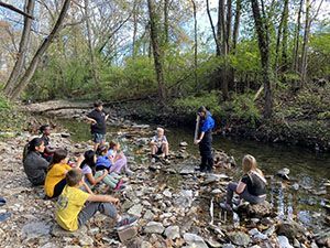 A group of people are sitting on rocks near a stream in the woods.