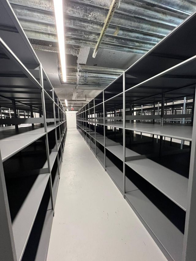 Empty warehouse aisle with tall black shelving on both sides and a white floor under overhead lights