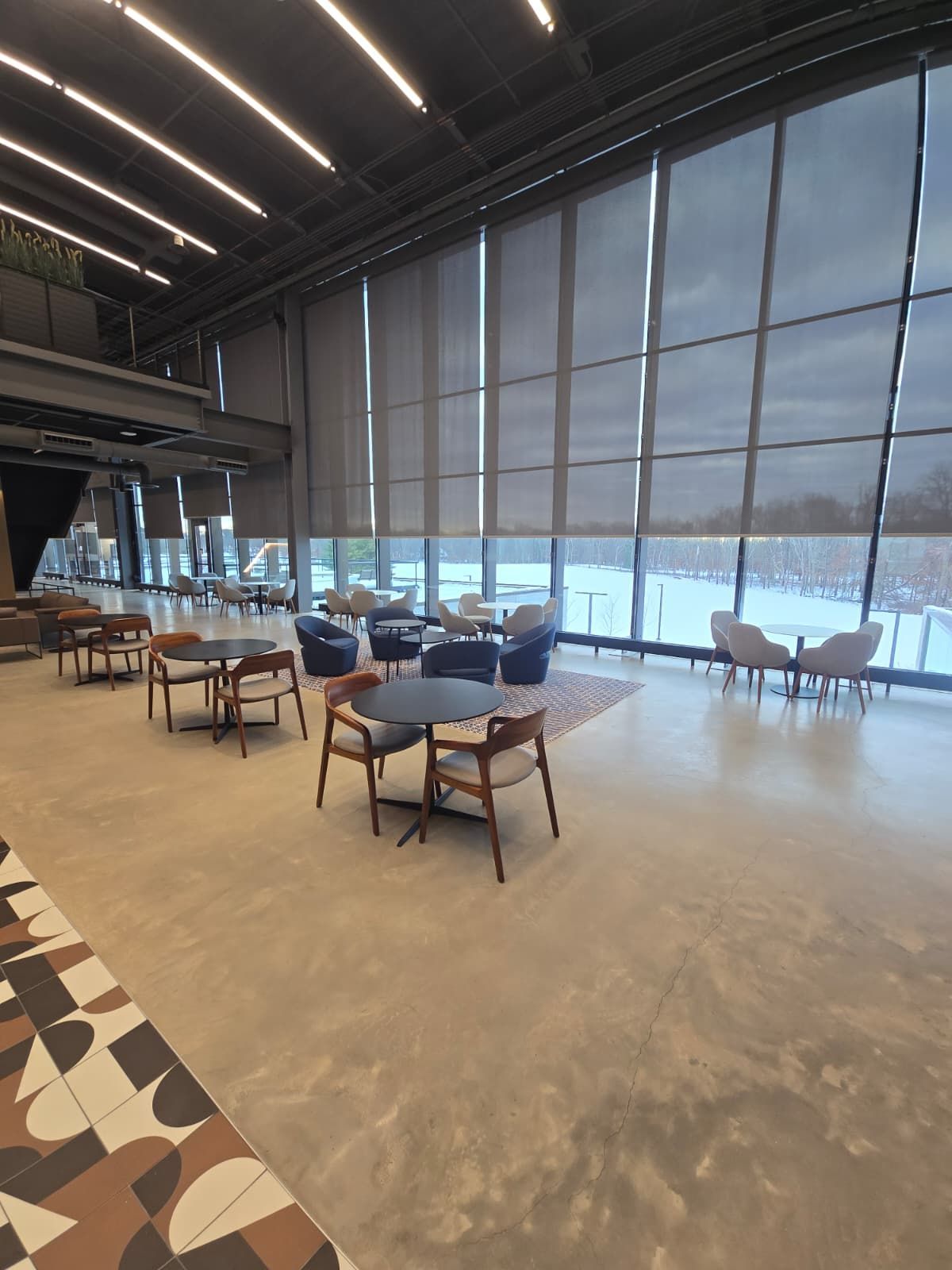 Modern lounge with blue chairs, wooden tables, patterned flooring, and floor-to-ceiling windows overlooking the water