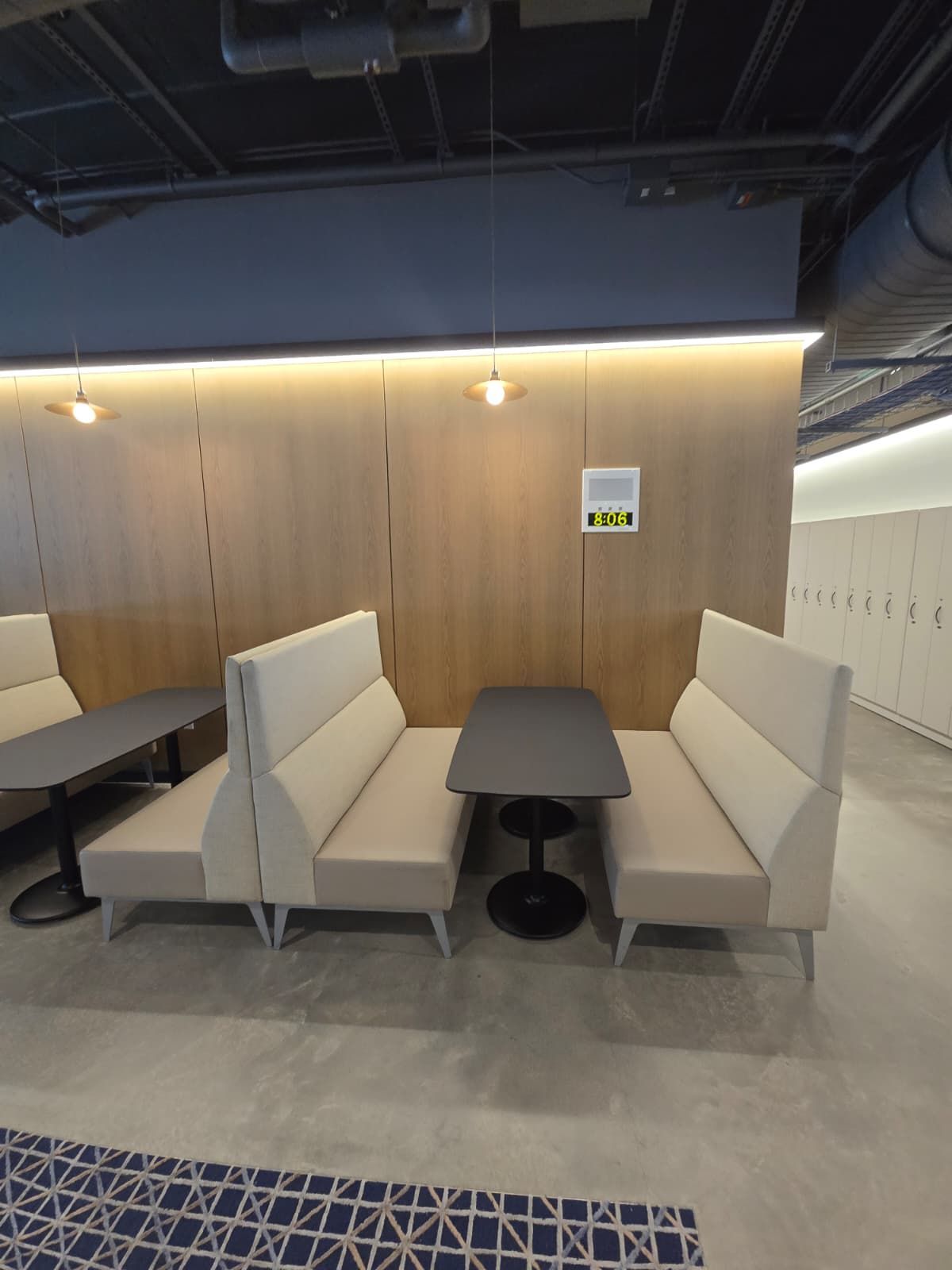 Modern lounge with beige booths around a dark table in a wood-paneled room, lit by a warm strip light.