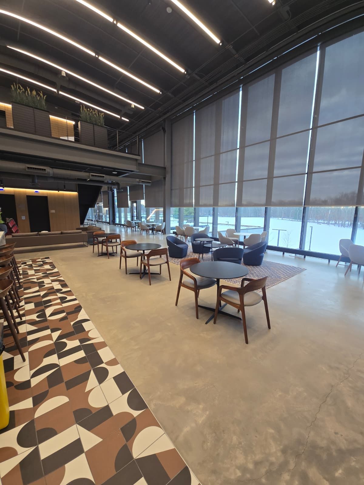 Modern lounge with chairs and tables, patterned floor, and large floor-to-ceiling windows overlooking the outdoors