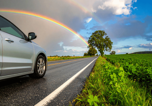 Gray car driving on road, with one side of the road one side stormy with rainbow another with green grass | Crowne Automotive