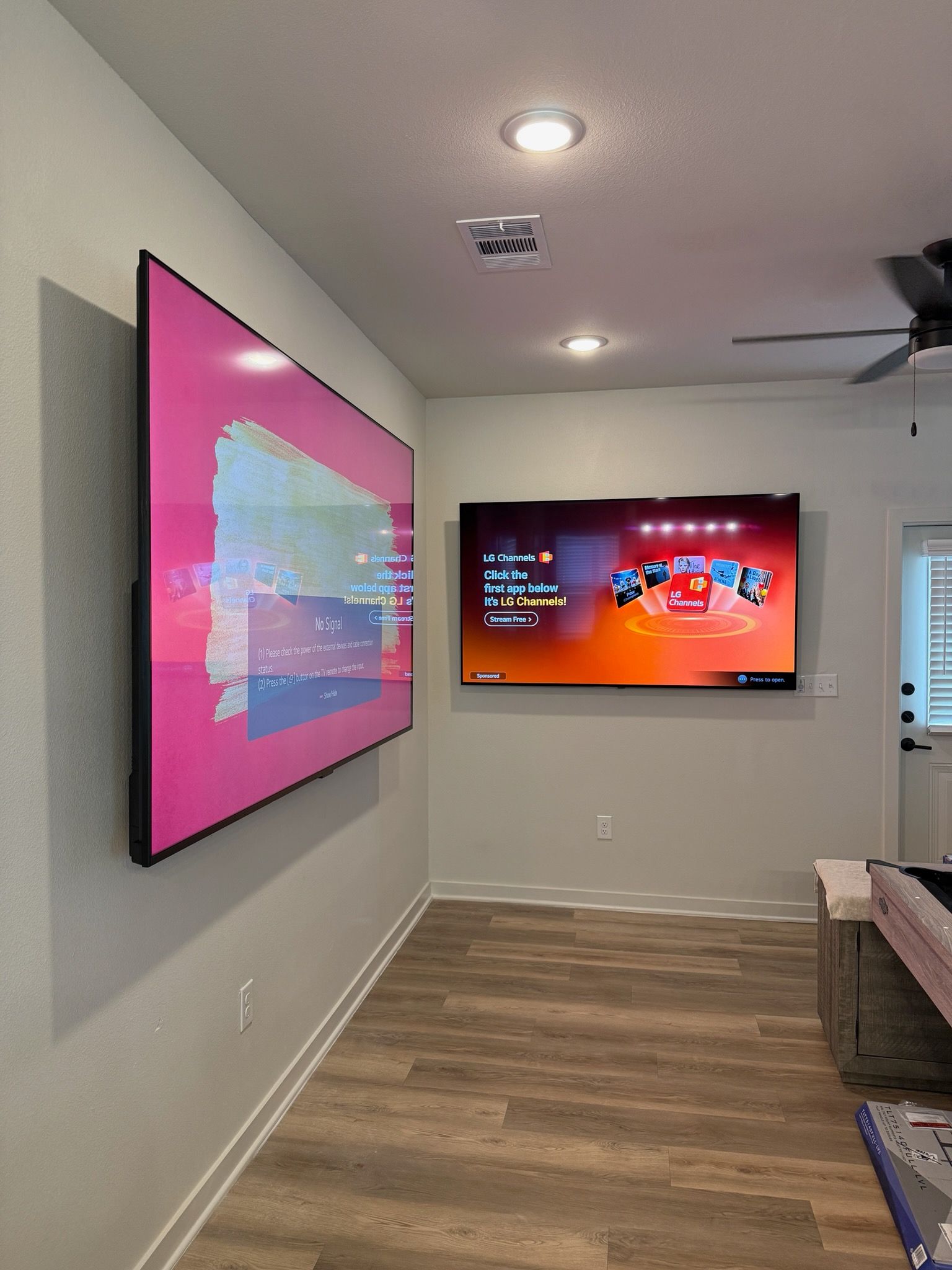 Two large TVs mounted on a light gray wall in a living room; one pink, one showing a game screen.