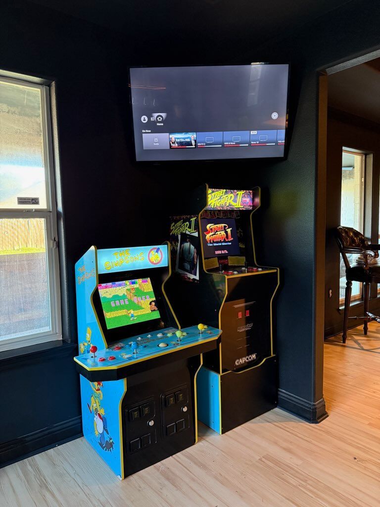 Arcade game machines beneath a mounted TV on a dark wall next to a window and doorway.